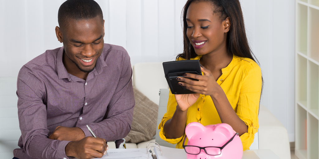 Happy Young Couple Calculating Bill With Piggybank On Desk
